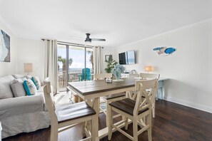 A spacious living area with a wooden dining table, white cushioned chairs, a sofa with blue and white cushions, and a large window offering a view of palm trees and the ocean. - Seaside Beach & Racquet 3205 (Orange Beach)