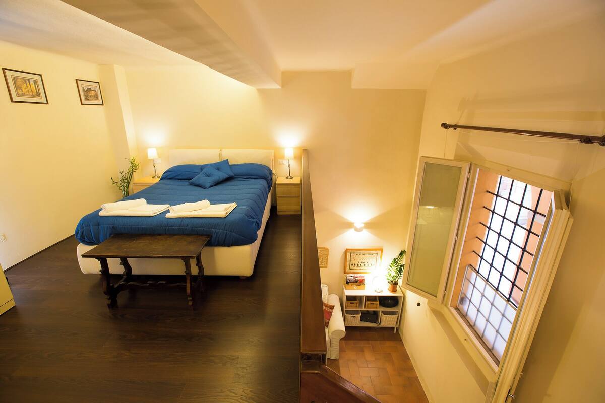 Un dormitorio con cama, mesitas de noche, un banco, una pequeña mesa, una ventana con barrotes y un estante fijado a la pared.
