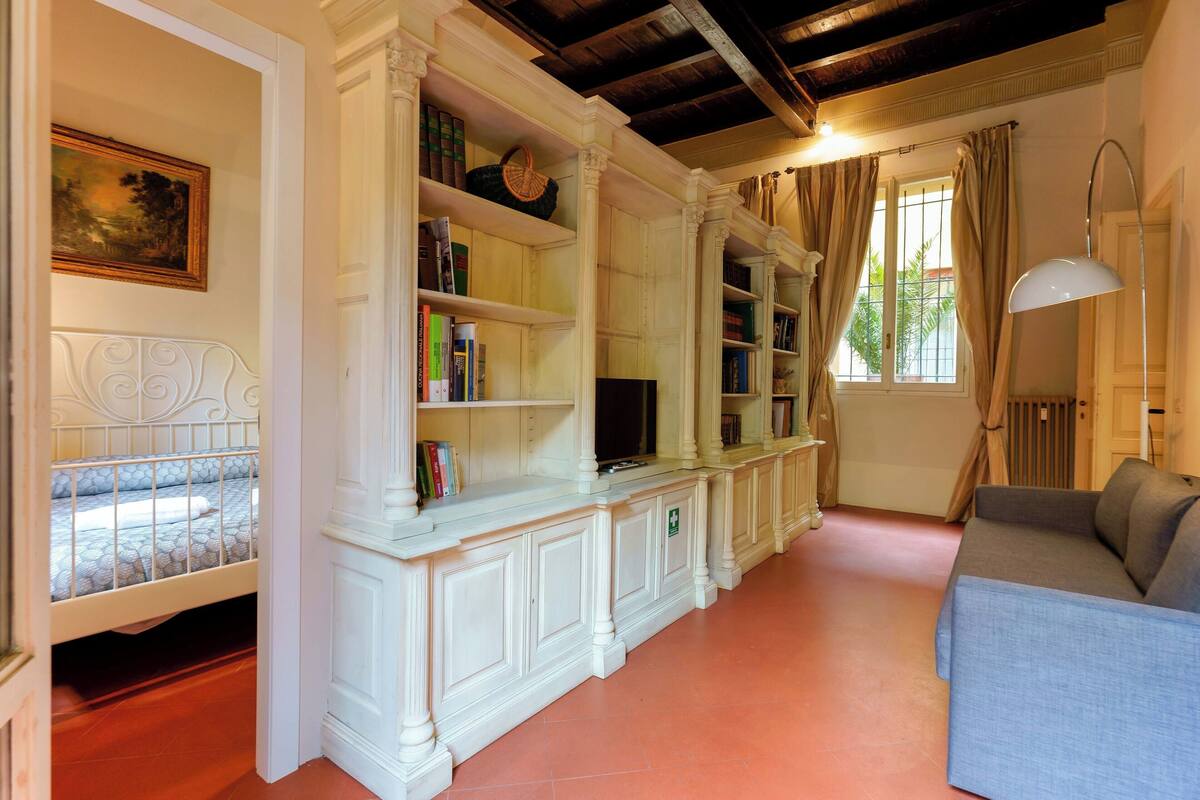 Una habitación con cama, sofá, estantería y ventana con cortinas.