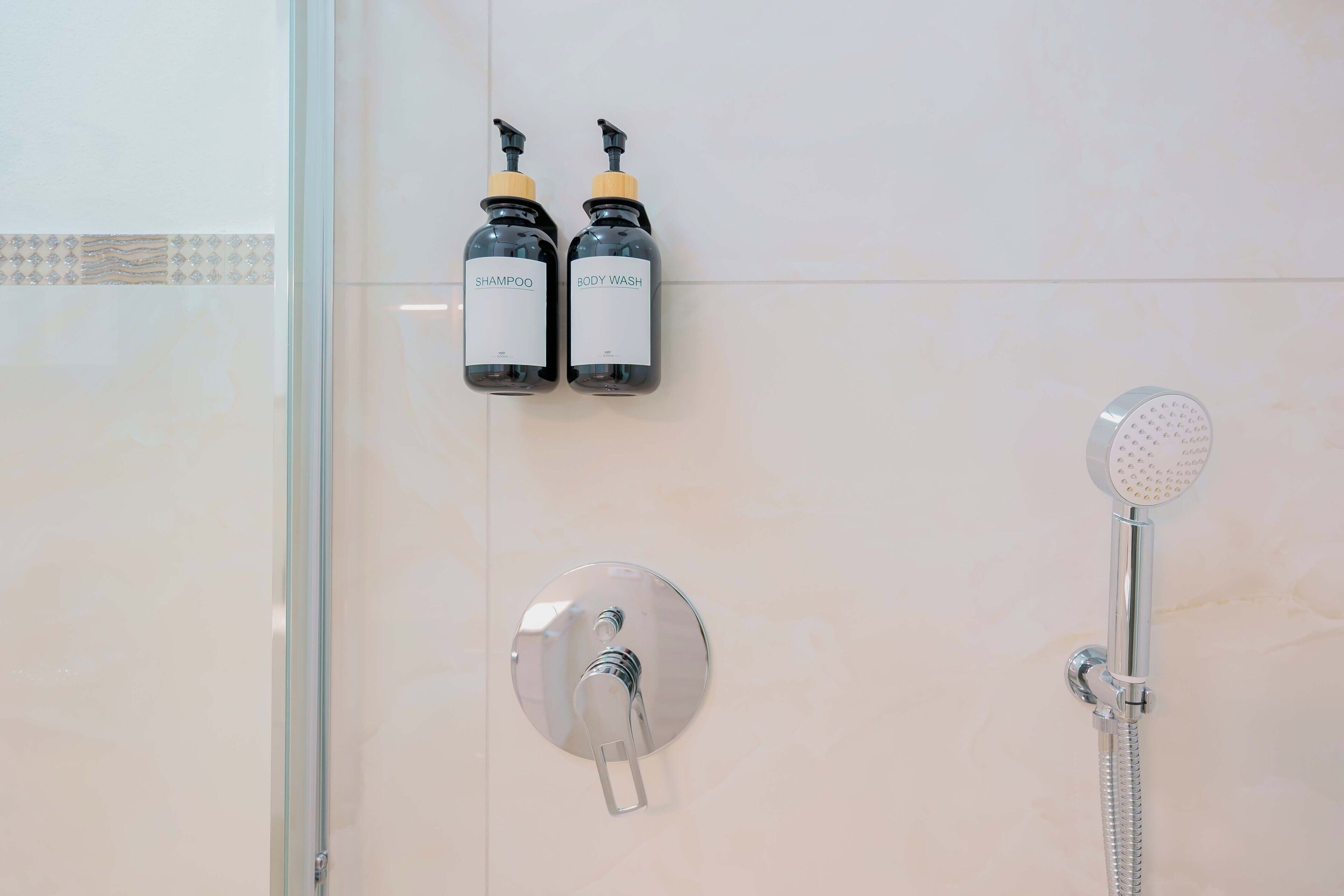 Et badeværelse med en bruser, to flasker mærket 'SHAMPOO' og 'BODY WASH', samt et vægmonteret armatur.
