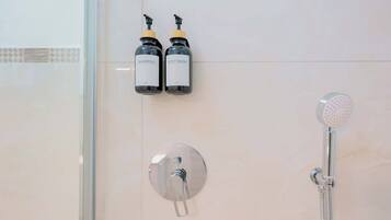 Et badeværelse med en bruser, to flasker mærket 'SHAMPOO' og 'BODY WASH', samt et vægmonteret armatur.