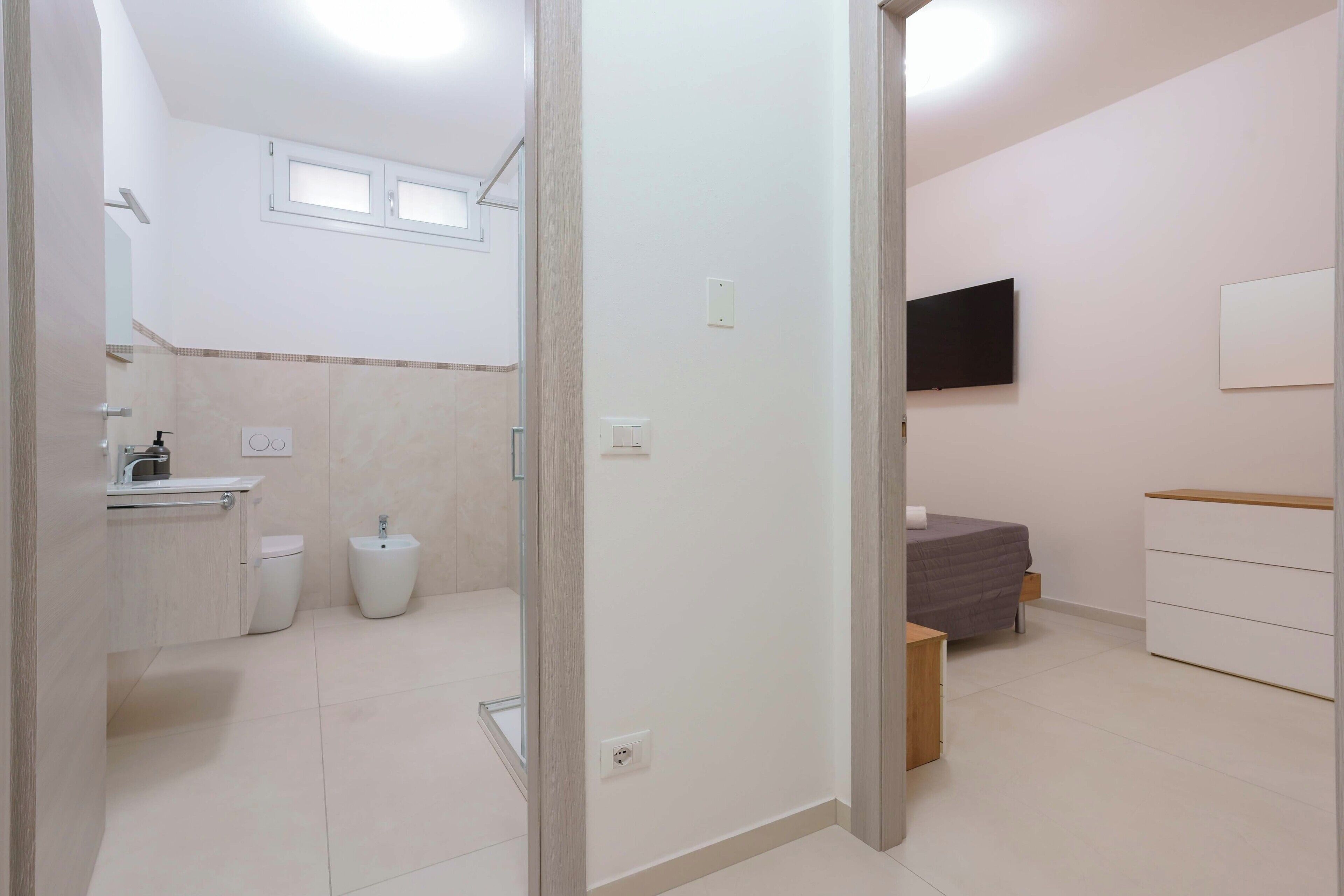 A modern bathroom with a toilet, bidet, and sink. There is a bed with a gray blanket and a TV mounted on the wall.
