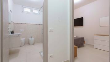 A modern bathroom with a toilet, bidet, and sink. There is a bed with a gray blanket and a TV mounted on the wall.
