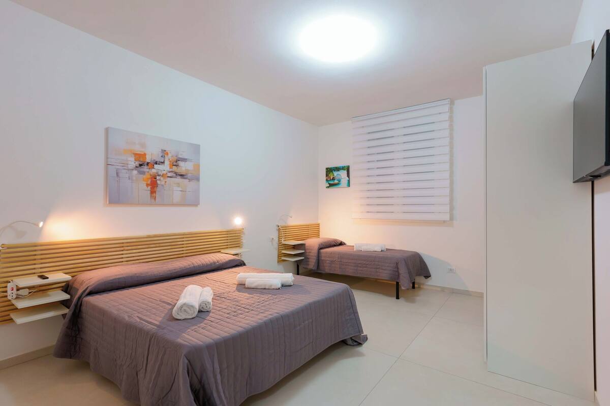 A hotel room with two beds, a flat-screen TV, a painting on the wall, and a window with blinds.