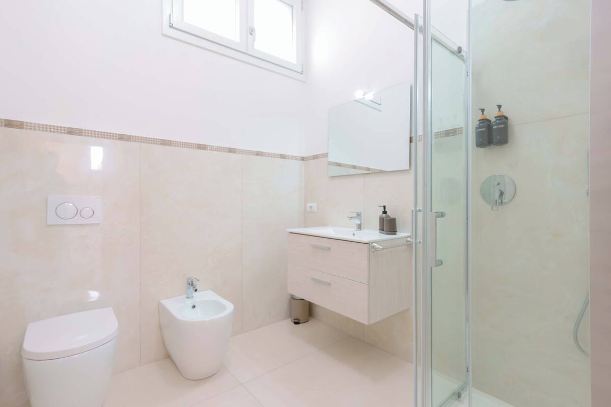 A modern bathroom with a toilet, bidet, vanity, mirror, and glass shower enclosure.