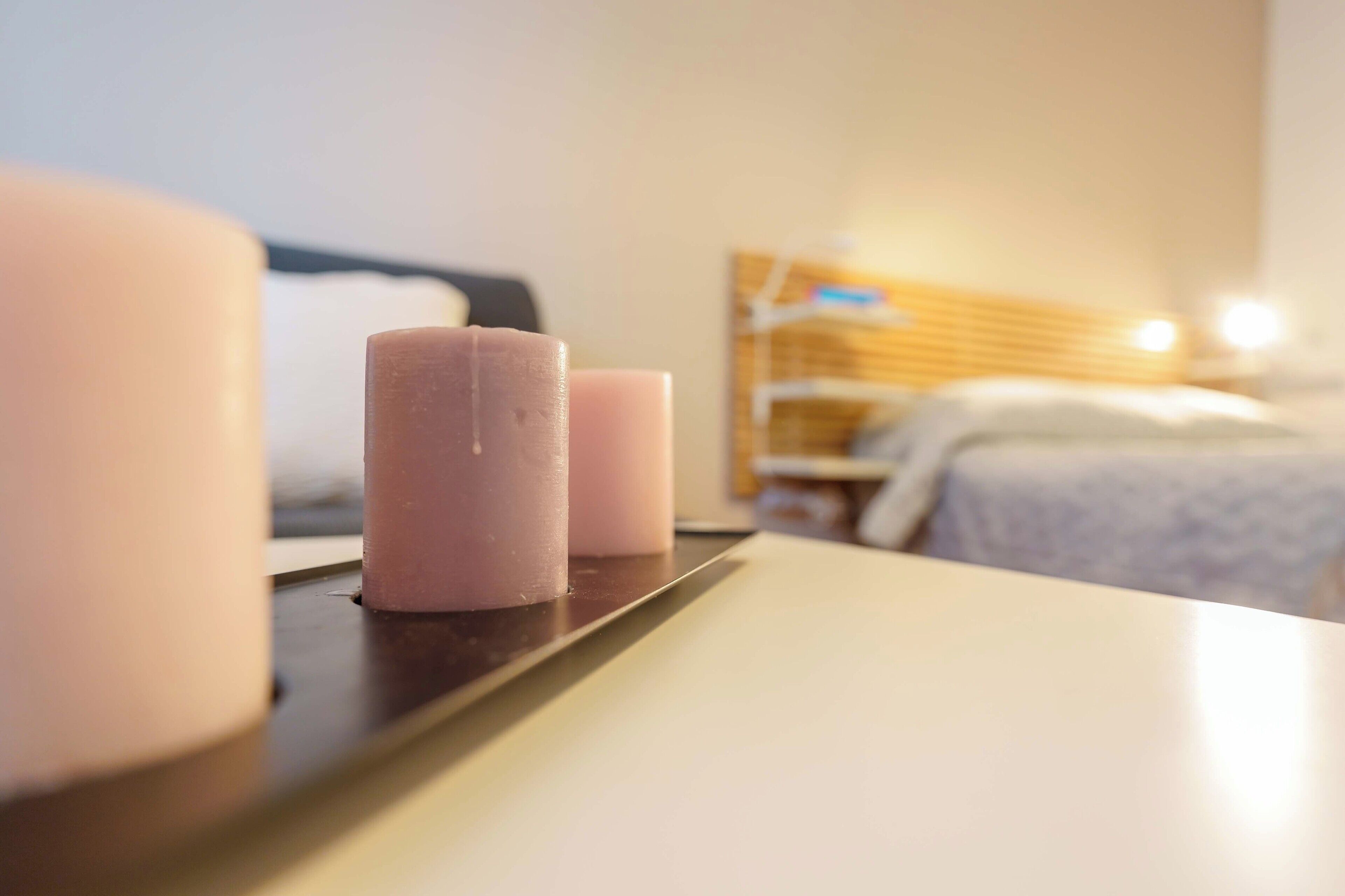 Primer plano de una mesa con tres velas. Al fondo, se ve una cama con cabecera de madera y una mesita de noche con una lámpara.
