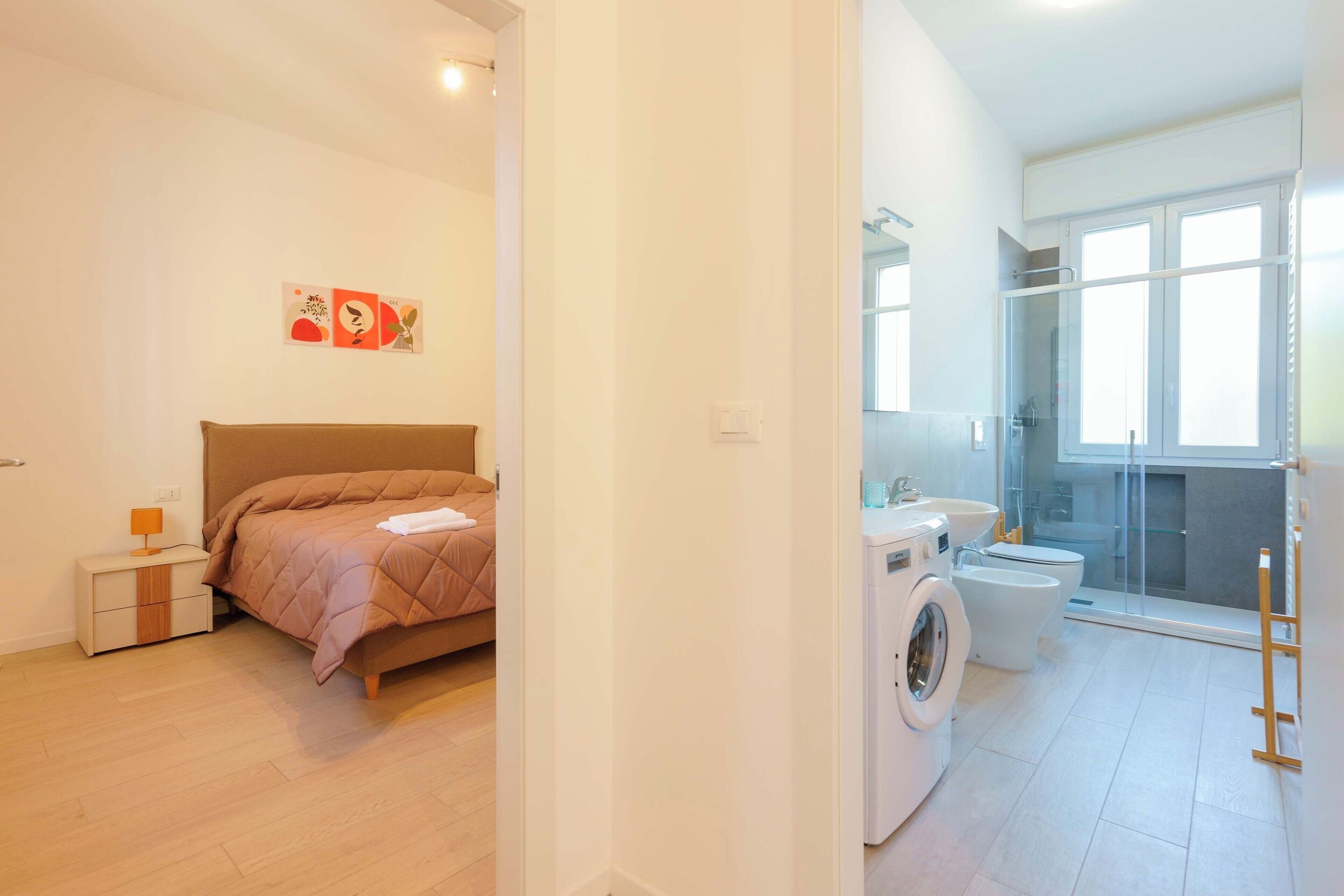Un dormitorio con cama, mesita de noche y un cuadro colgado en la pared.  Cerca, un baño con lavadora, inodoro y cabina de ducha de vidrio.