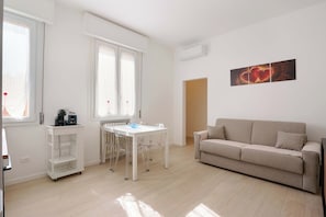 Een moderne woonkamer met een beige bank, een witte eettafel met stoelen en een koffiezetapparaat op een witte trolley.