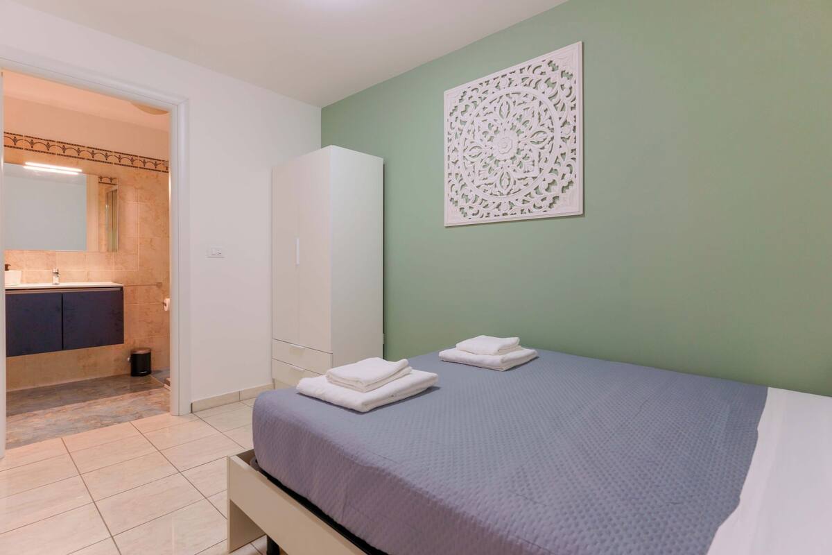 Een slaapkamer met een bed, een badkamer met een wastafel en spiegel, en een groene muur met een decoratief paneel.