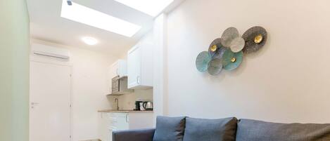 Una sala de estar moderna con un sofá gris, una zona de cocina con armarios blancos y un elemento decorativo en la pared.