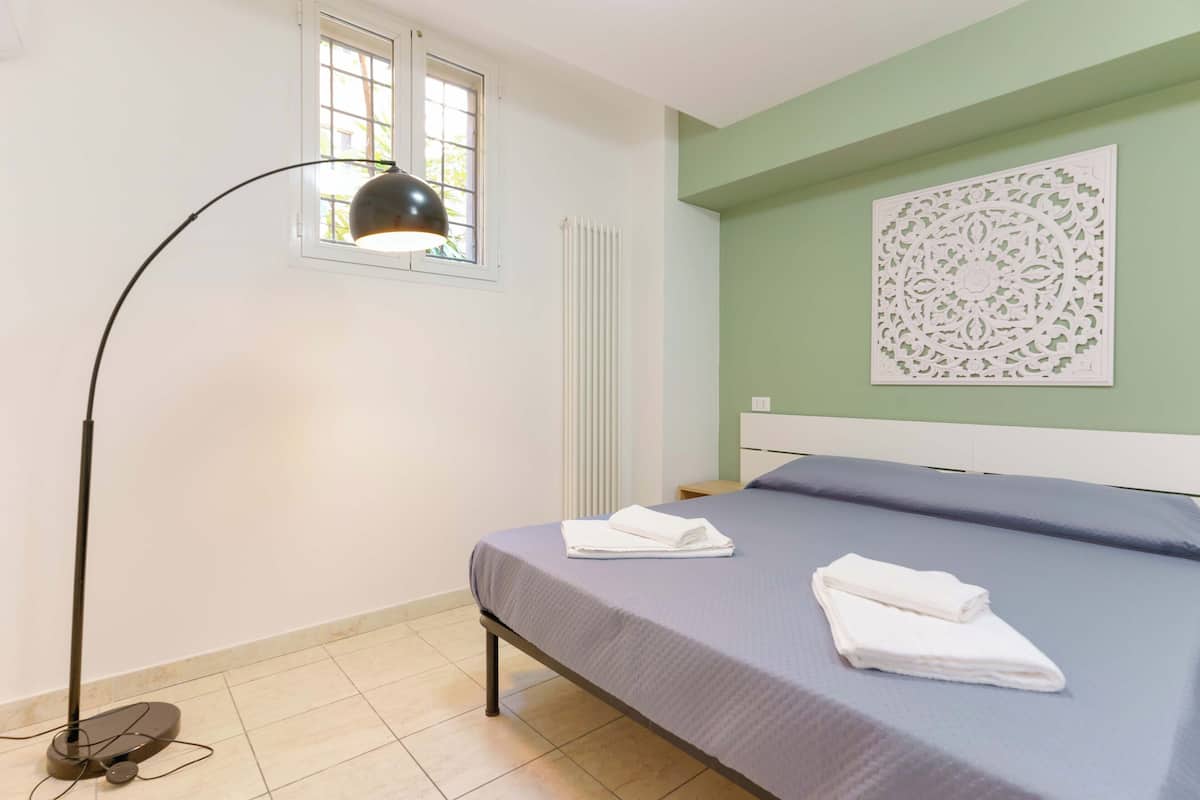 Een slaapkamer met een bed, een vloerlamp, een radiator, een raam en een decoratief wandobject.