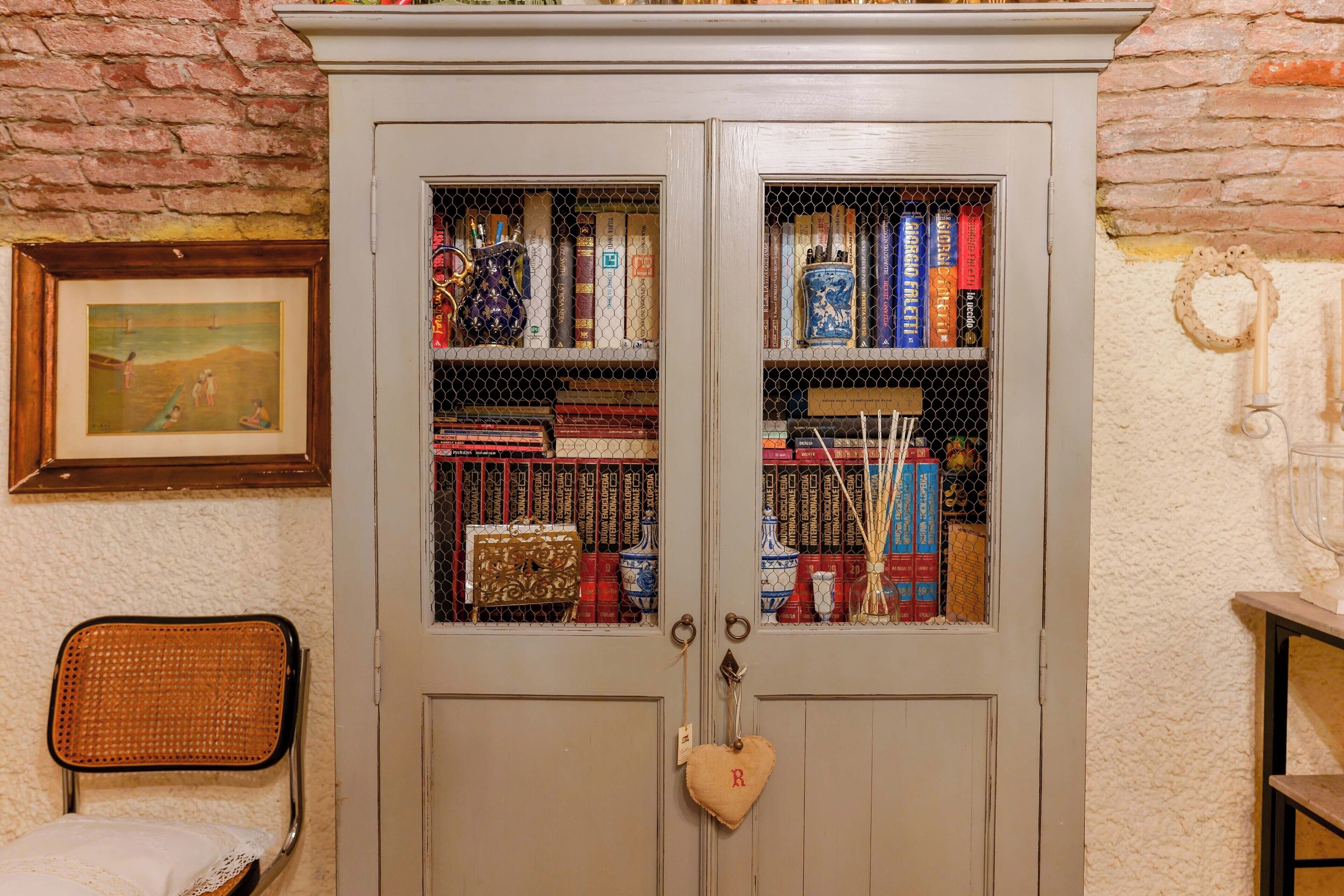 Una habitació amb una biblioteca plena de llibres i objectes decoratius, una cadira de vimet i un quadre encapçalat a la paret.