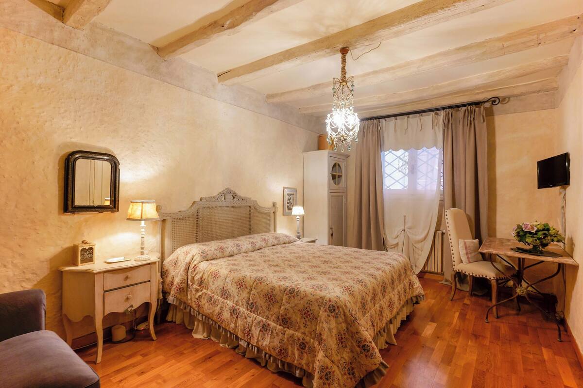 Une chambre avec un plancher en bois, un lit recouvert d’une couvre-lit à motifs floraux, une table de chevet avec une lampe, une commode, une chaise et un bureau agrémenté d’un vase de fleurs.