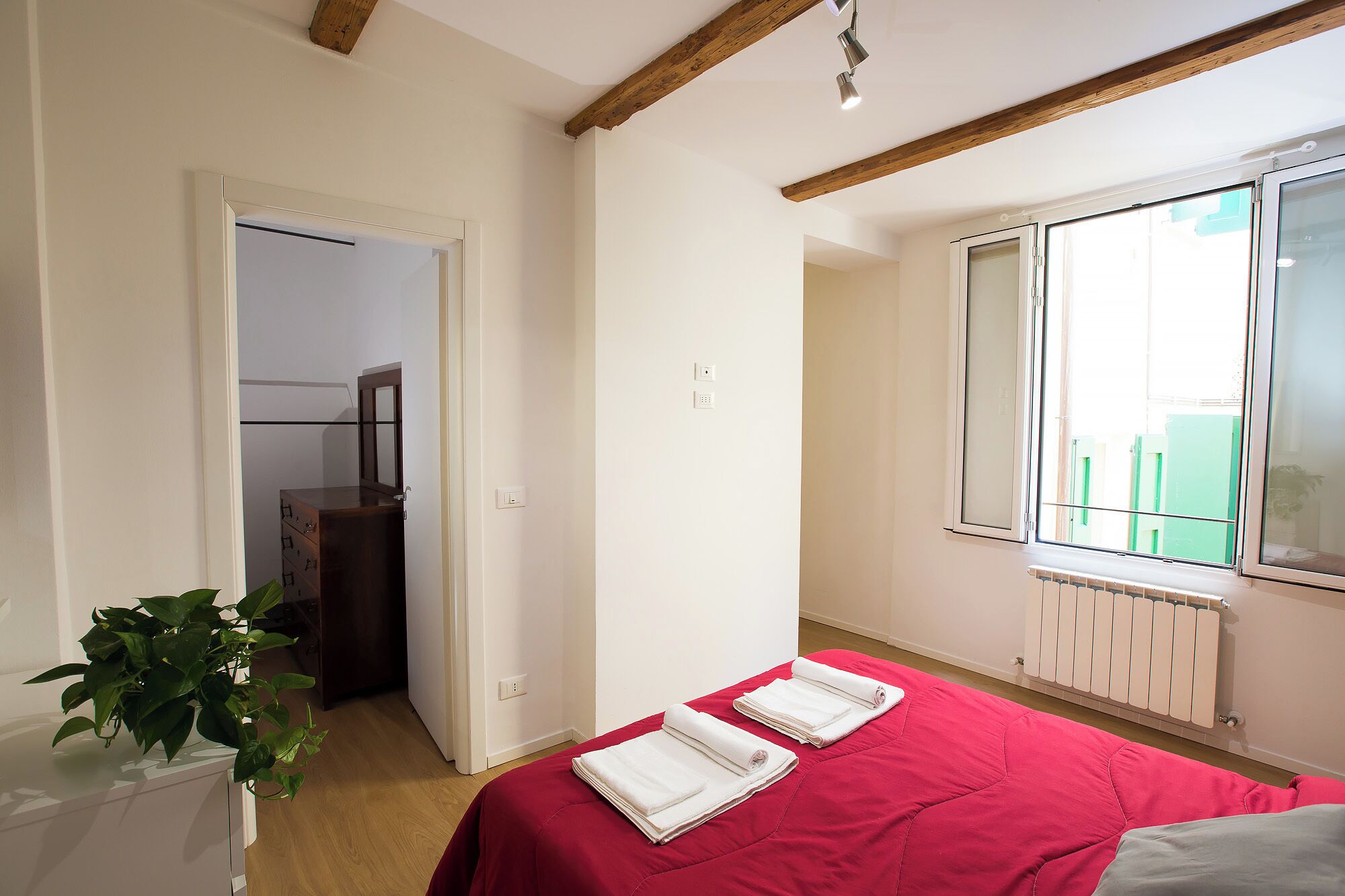 A bedroom with a red bedspread, a wooden ceiling, a window with a view, and a plant on a dresser.