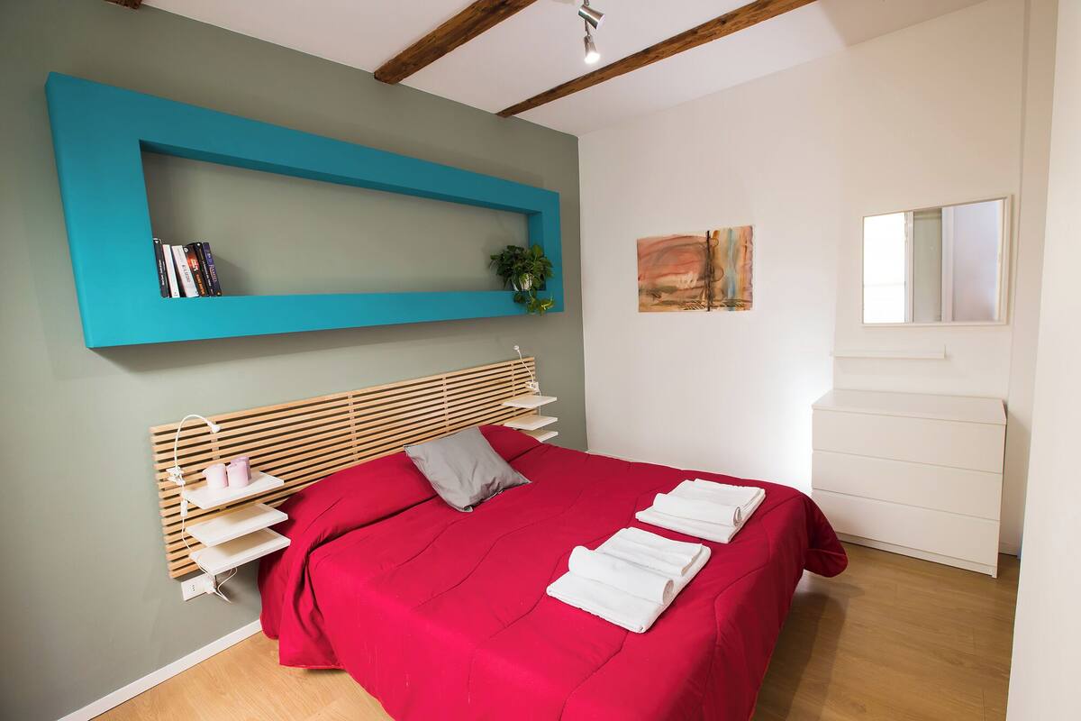 A bedroom with a bed, a bookshelf, a plant, and a painting on the wall.