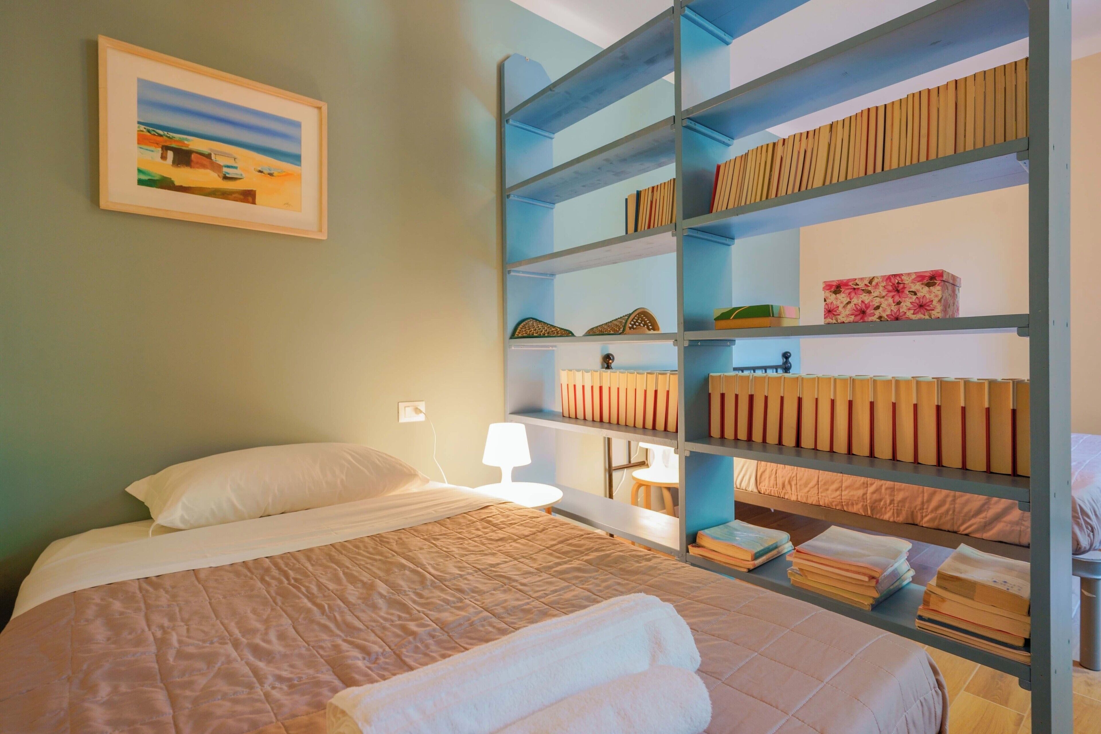 Een slaapkamer met een bed, nachtkastje, lamp, boekenplank en een schilderij aan de muur.