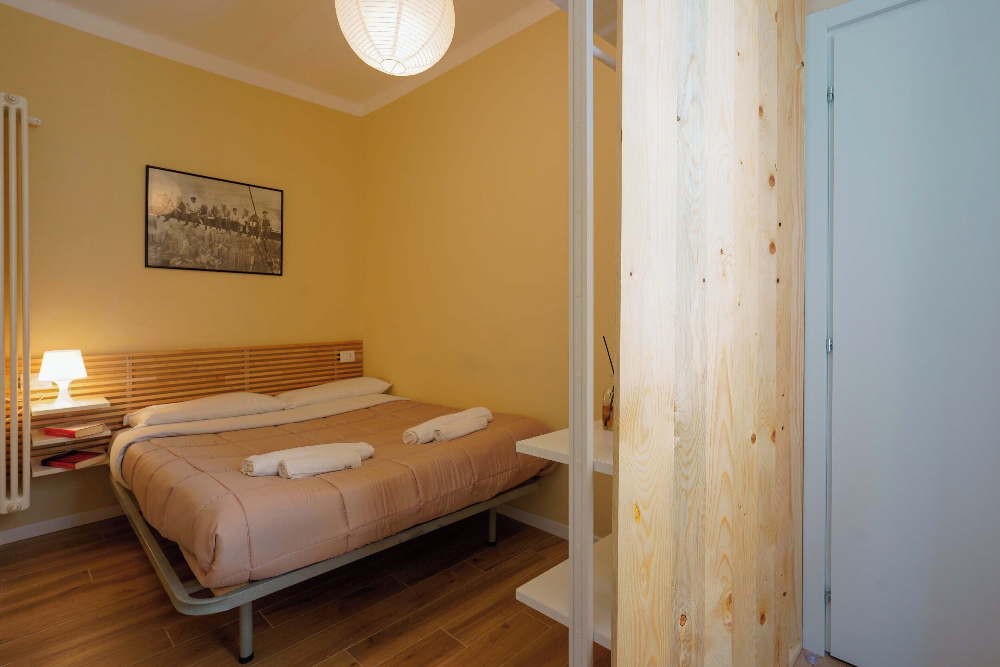 Een kamer met een bed, een nachtkastje met een lamp, een ingelijst schilderij aan de muur en een houten deur.