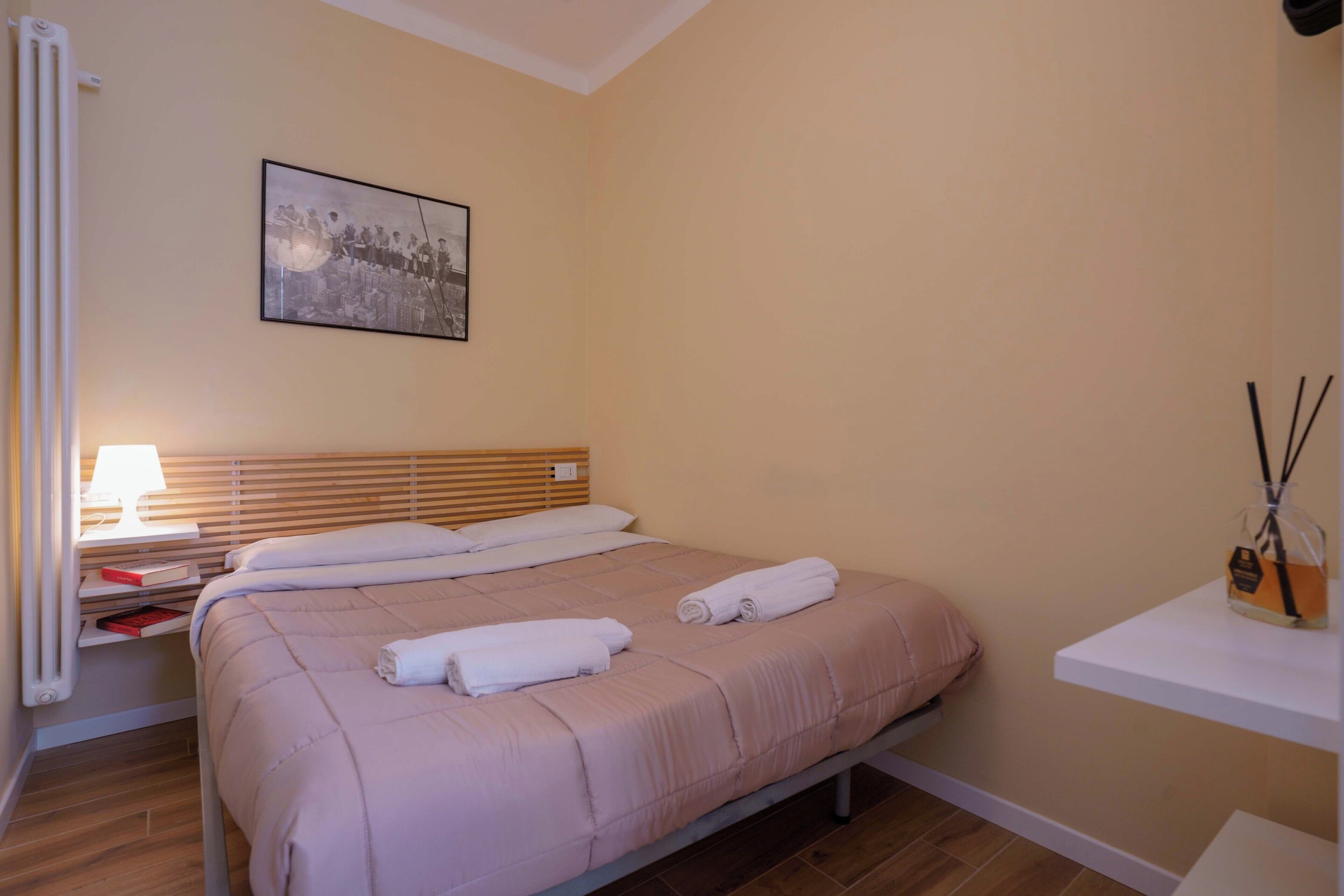 A hotel room with a bed, a nightstand, a lamp, a picture on the wall, and a shelf with a bottle.