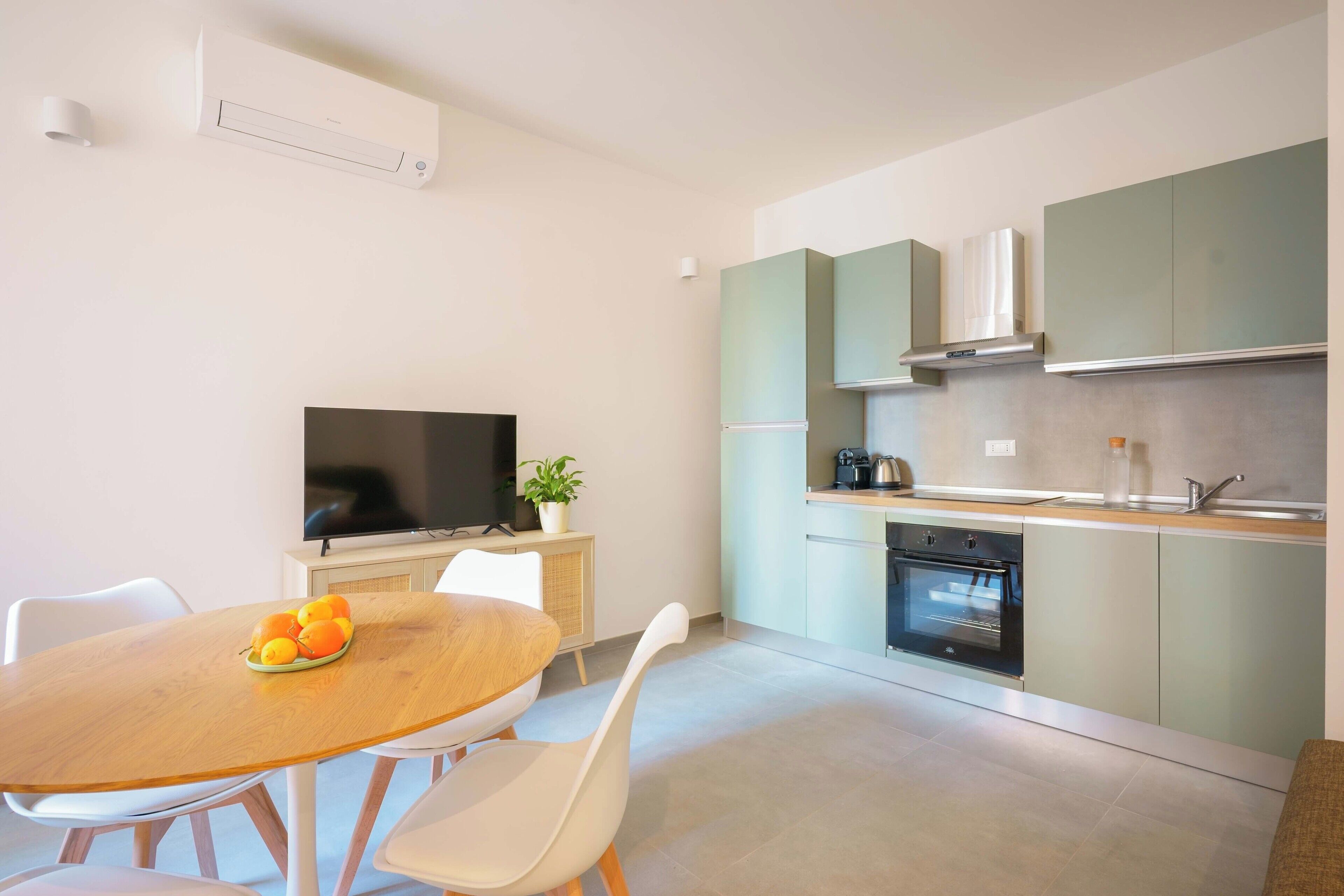 A modern kitchen with a dining area, a flat-screen TV, and a bowl of fruit on the table.