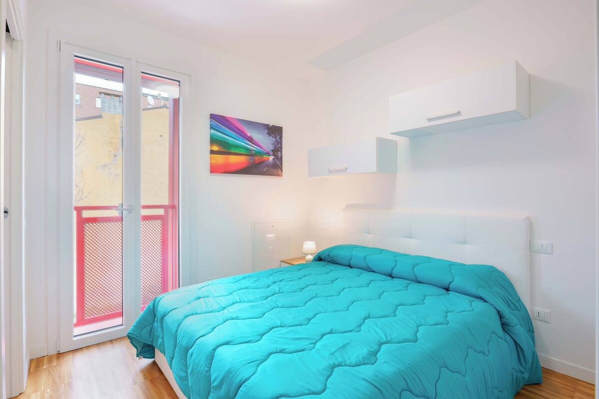 A bedroom with a bed, a window with a view, a painting on the wall, and a white cabinet.