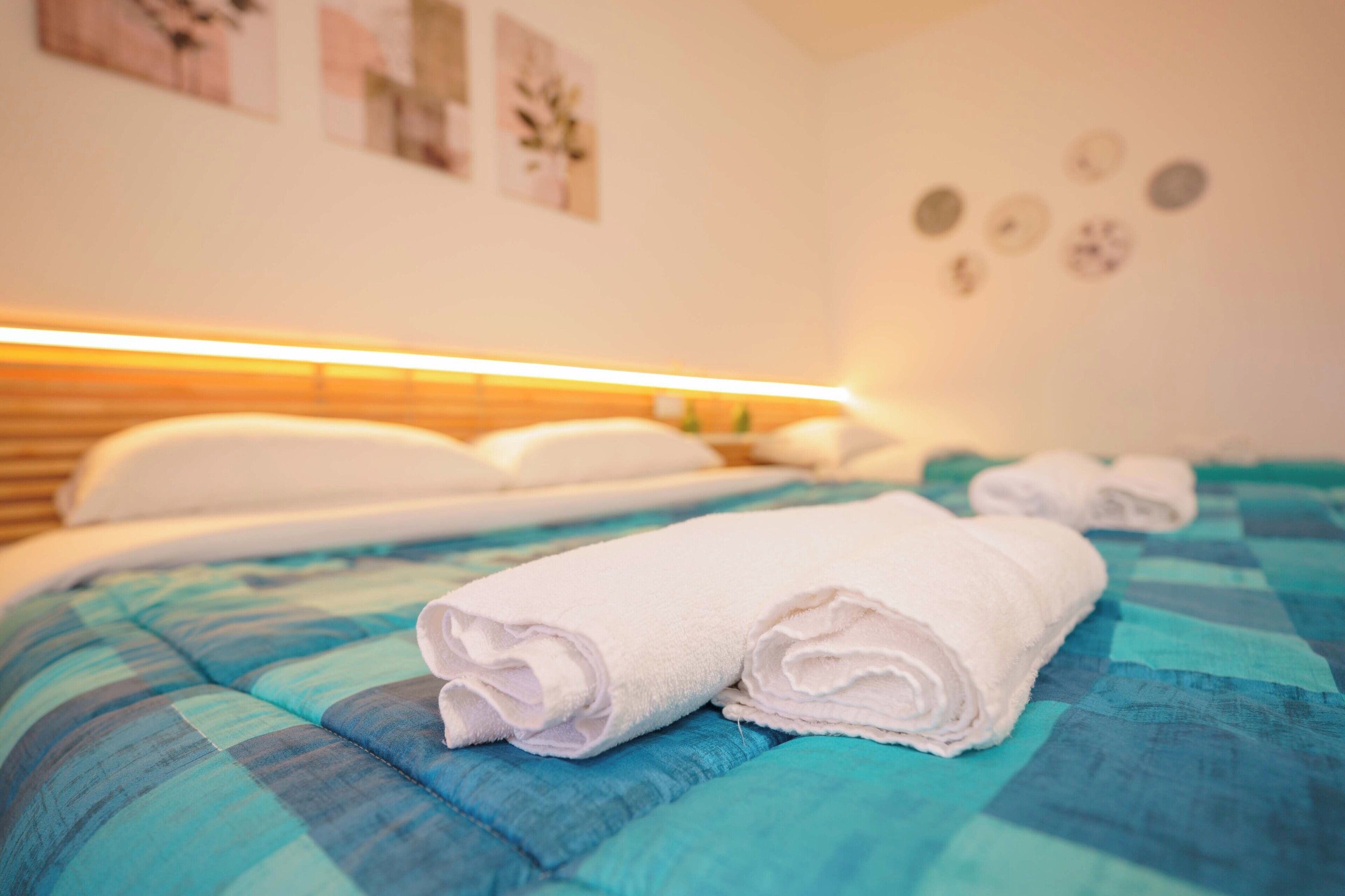 Habitación de hotel con cama, almohadas, colcha azul y verde azulada, y toallas blancas enrolladas.