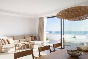Una sala de estar moderna con un sofá, sillas, una mesa de comedor y un ventanal con vista al mar.