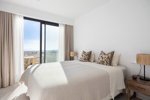 Habitación de hotel con una cama grande, dos lámparas de noche, una mesita de noche de madera y vistas de la ciudad a través de ventanas de piso a techo.