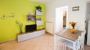 Una sala de estar moderna con una pared verde, un televisor de pantalla plana, una mesa de comedor con sillas y una planta pequeña en la pared.