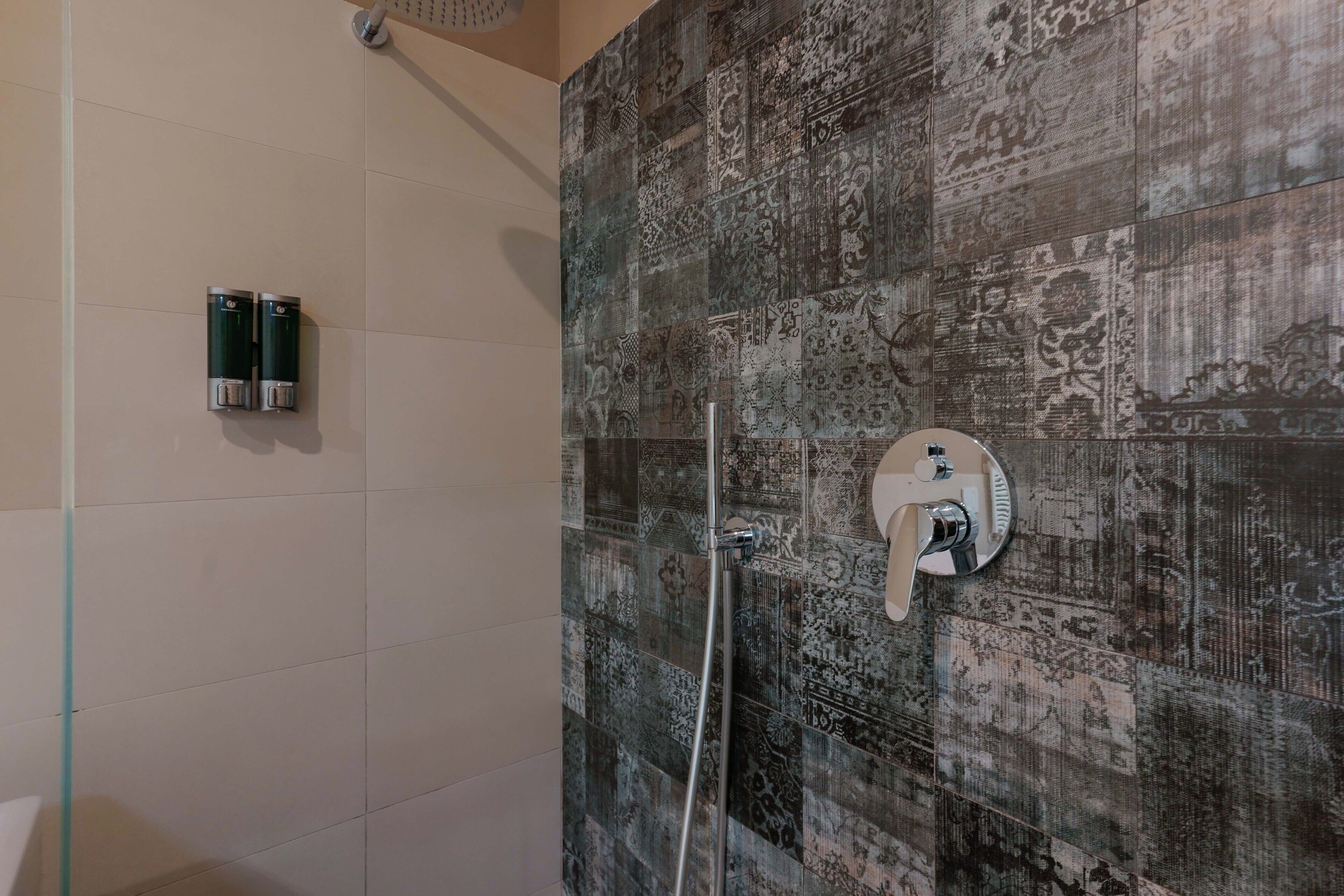 Un espacio de ducha con baldosas estampadas, un cabezal de ducha de mano y dos secadores de manos fijos en la pared.