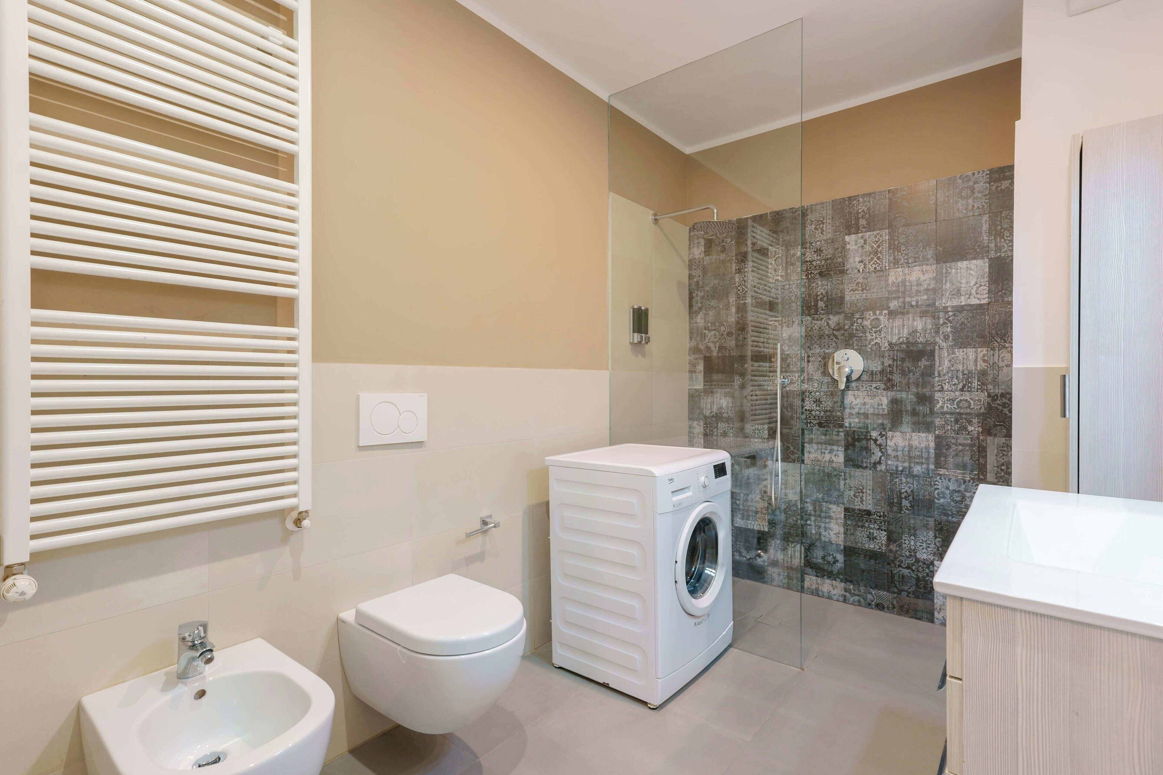 A modern bathroom with a bidet, toilet, washing machine, and a shower area with a patterned wall.