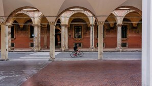 An open courtyard with arches, a person on a bicycle, and a brick building in the background. - Zamboni 58 Apartment (Bologna)