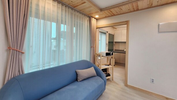 A modern room with a blue sofa, wooden ceiling, and a kitchen area visible through an open door. - Sokcho Family House Near Seorak Port, Seafood Market & Beach (Walkable) (Sokcho-si)