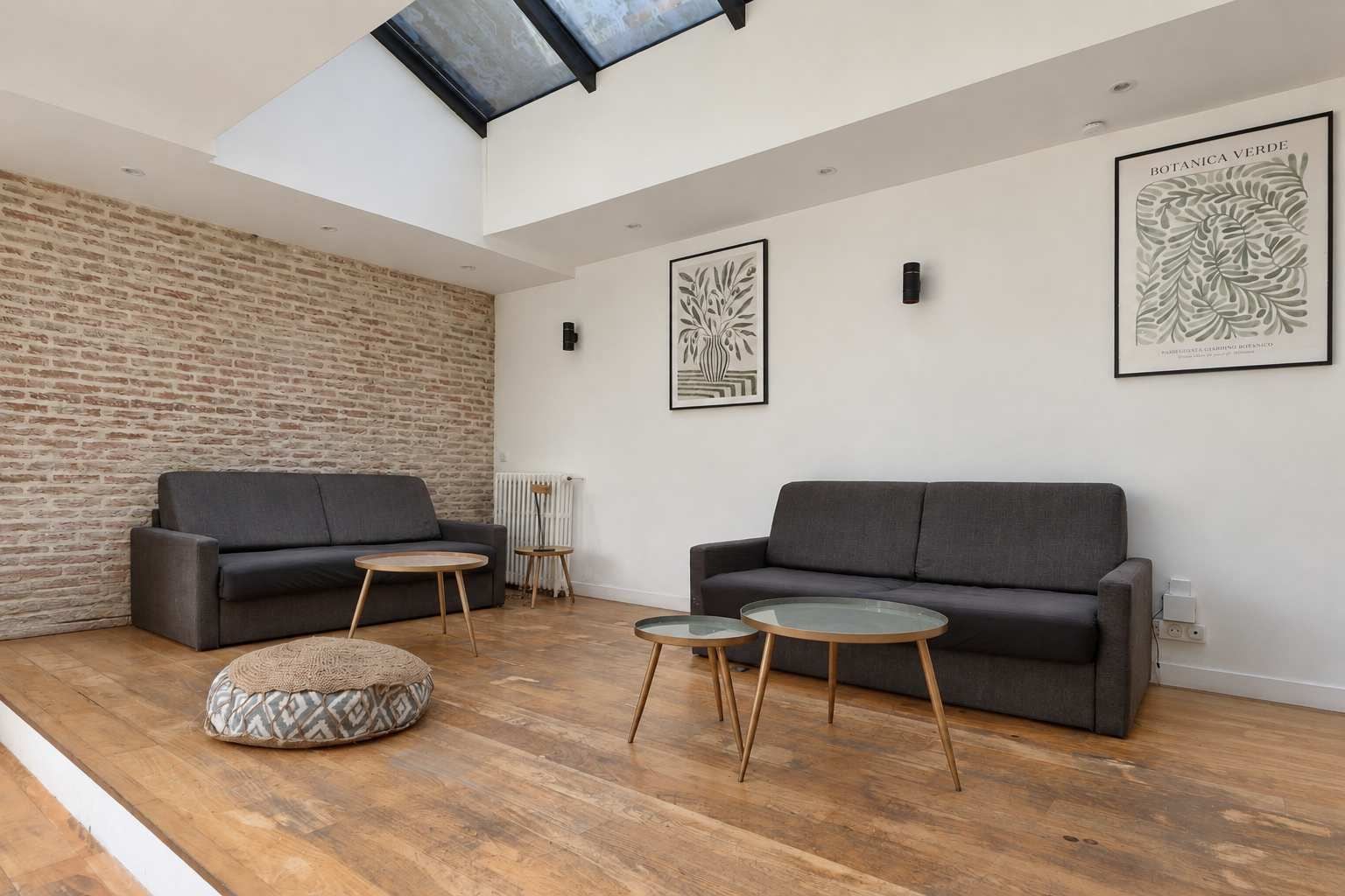 A modern living room with two sofas, round tables, a patterned ottoman, and a brick wall.