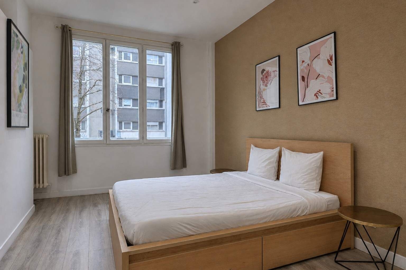 Une chambre à coucher avec un lit, une fenêtre avec des rideaux, un radiateur et deux tableaux encadrés accrochés au mur.