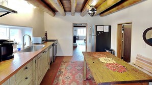 A kitchen with wooden cabinets, a sink, a stove, and a refrigerator. There is a rug on the floor and a table with a floral design. - Casa Sarita (Santa Fe)