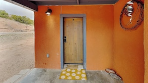 A wooden door with a metal handle, a yellow patterned mat, and a wall-mounted light fixture. - Casa Sarita (Santa Fe)