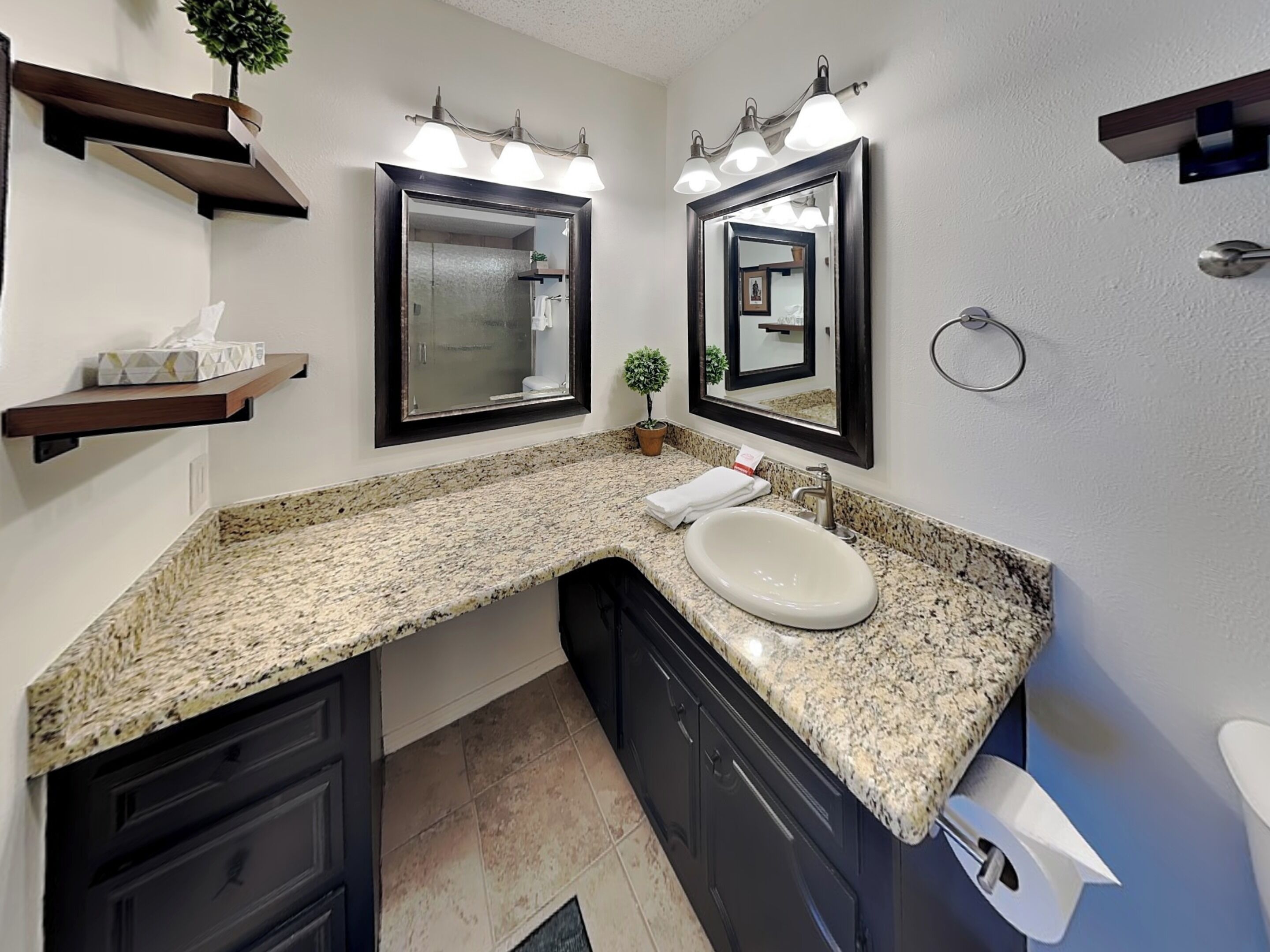 Une salle de bain dotée d’un comptoir en granit, d’un lavabo blanc, de meubles sombres et de deux miroirs fixés au mur, chacun agrémenté d’un luminaire.