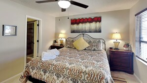 Une chambre à coucher avec un lit, deux tables de chevet avec des lampes, un ventilateur de plafond, un tableau au mur et une porte donnant sur une salle de bain.