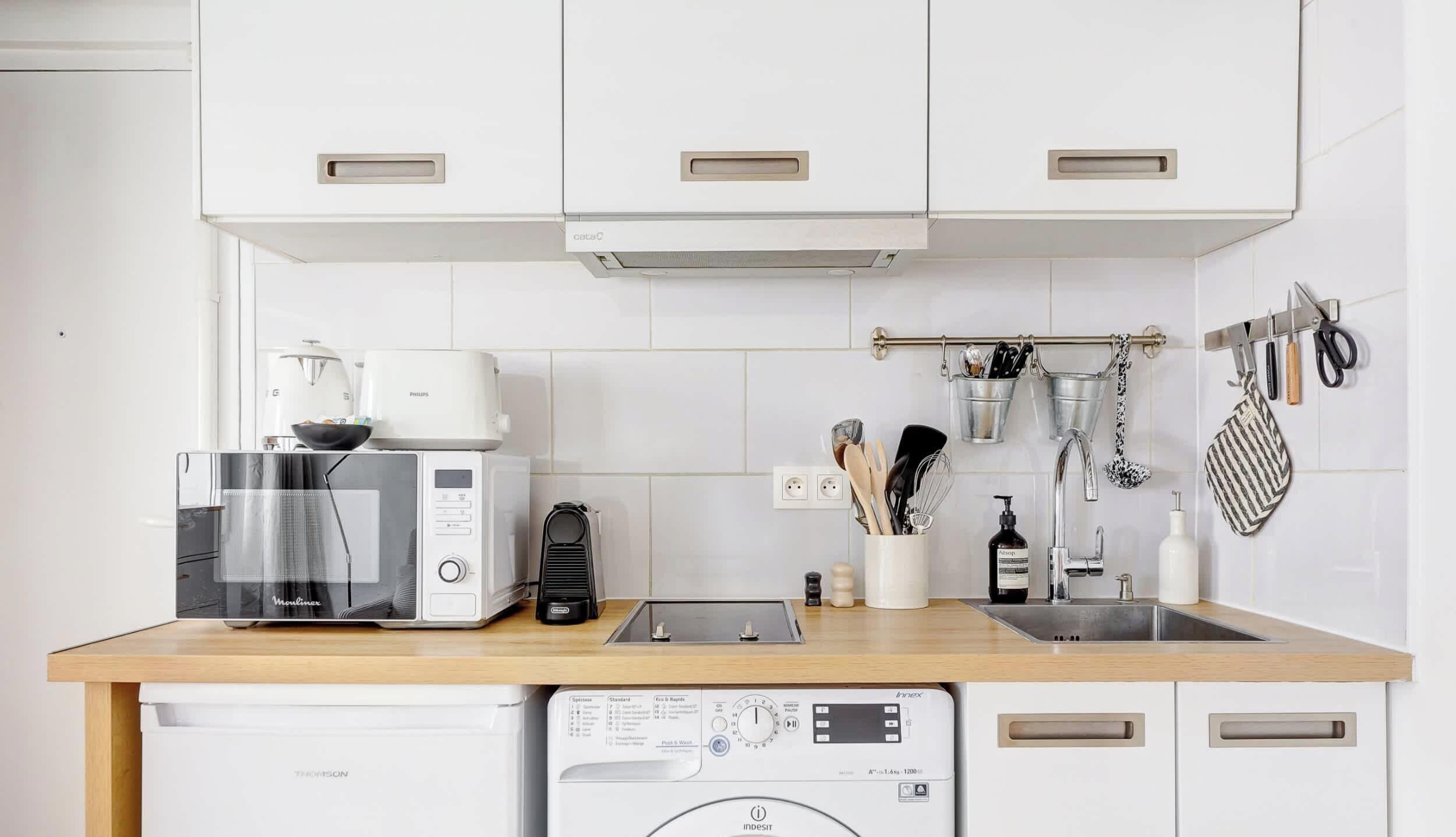 Une cuisine compacte équipée d’un four à micro-ondes, d’un grille-pain et d’un lave-linge. Des ustensiles sont accrochés au mur, et il y a un évier avec un robinet.