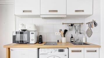 Une cuisine compacte équipée d’un four à micro-ondes, d’un grille-pain et d’un lave-linge. Des ustensiles sont accrochés au mur, et il y a un évier avec un robinet.