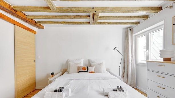 Una camera da letto con soffitto in legno, un letto con lenzuola bianche, un comodino con una lampada e una finestra con tende.