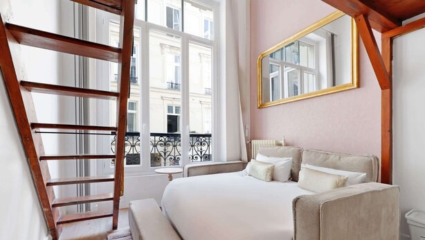 Una camera da letto con una scala in legno, un letto con biancheria bianca, uno specchio grande e vista sugli edifici attraverso una finestra.