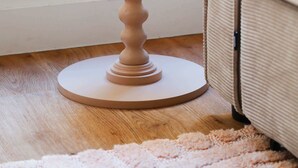 Un tavolo rotondo in legno con base a colonna, un divano beige e un tappeto rosa in una stanza con pavimento in legno e una finestra con inferriate decorative.