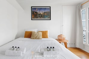 A hotel room with a bed, white bedding, a framed painting, a small table with a vase, and a window with curtains. - Studio elegant pres de Saint-Sulpice (Paris)