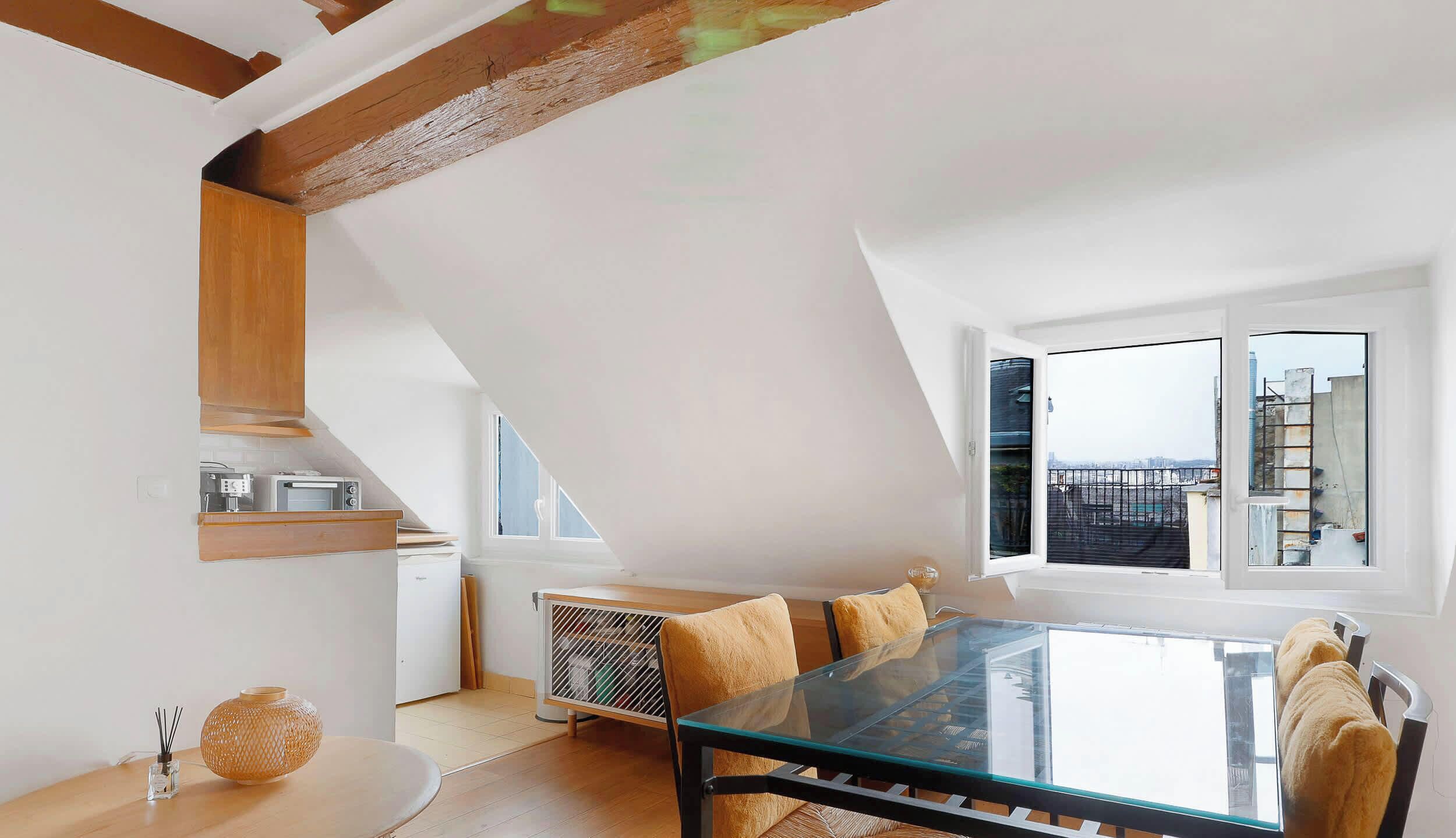 A modern loft apartment with a dining area featuring a glass table and wooden chairs, a kitchen with built-in appliances, and a large window offering a city view.