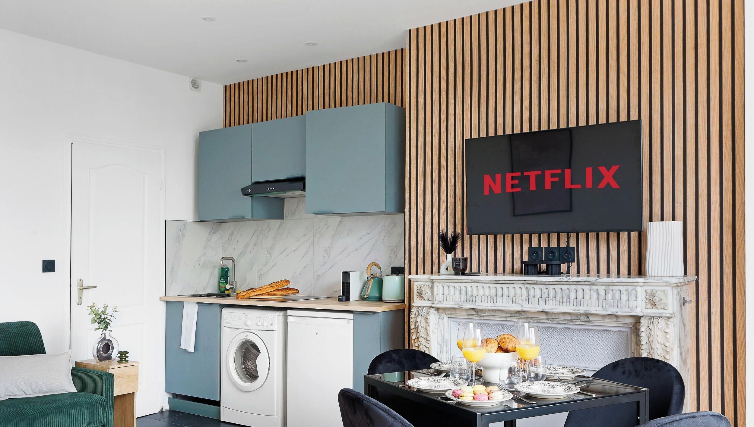 Ett modernt kök med en inbyggd tvättmaskin, en matplats med frukostmöblemang och en väggmonterad TV som visar ”NETFLIX”.