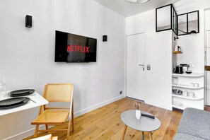 Ein modernes Wohnzimmer mit einem an der Wand befestigten Flachbildfernseher, einem Essbereich mit einem weißen Tisch und Stühlen sowie einer Küche mit weißen Schränken und Regalen.