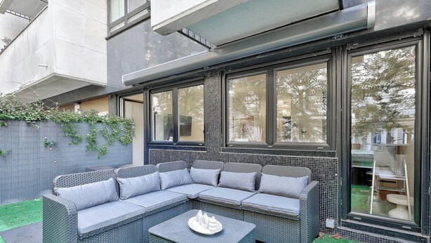 A modern outdoor seating area with a sectional sofa and a small table, surrounded by large windows and a metal fence. - BM Magnifique Studio avec terrasse - Arc de Triomphe (Paris)