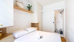 Una habitación de hotel moderna con una cama grande, una mesita de noche, un estante de madera con una planta y un baño visible a través de una puerta abierta.