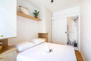 A modern hotel room with a large bed, a bedside table, a wooden shelf with a plant, and a bathroom visible through an open door. - Appartement 2 pièces à proximité de Gare de L'est (Paris)